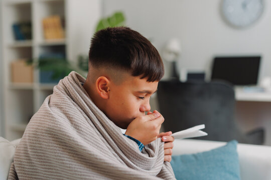 Young boy bundled in a blanket, coughing into his hand and holding a tissue while resting at home, conveying cold symptoms, childhood illness, and the need for care