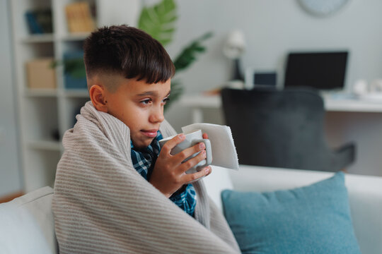 Young boy wrapped in a blanket, holding a cup of tea and resting on a sofa at home, finding comfort and warmth while recovering from a cold or flu during winter
