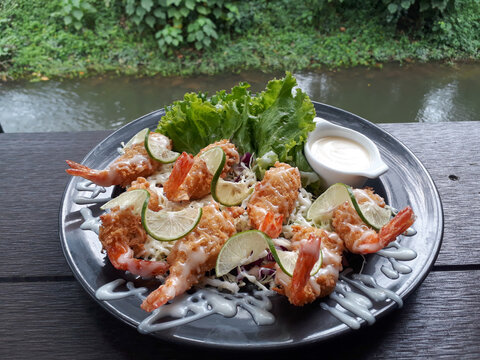 Delicious deep-fried breaded shrimp topped with creamy dressing and fresh lime slices, served with mixed salad on a dark plate over a rustic wooden table with a scenic natural river view. Perfect for 