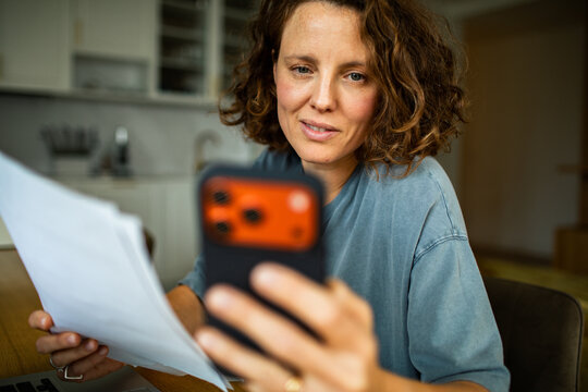 Woman reviewing paperwork and checking smartphone at kitchen table
