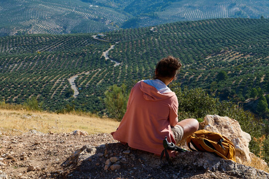 Mountain hiking rest with a hiker seated on a rock, looking across a wide valley landscape with trekking poles and backpack