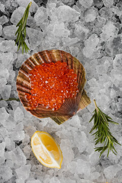 Red salmon roe served in scallop shell on crushed ice, accompanied by lemon wedge and sprigs of rosemary. Top-down view; bright orange roe glistens against gray background. Luxurious seafood appetizer