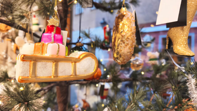 Colorful glass Christmas ornament shaped like a festive gift on a golden sleigh, hanging on a decorated Christmas tree with sparkling lights and seasonal decorations.