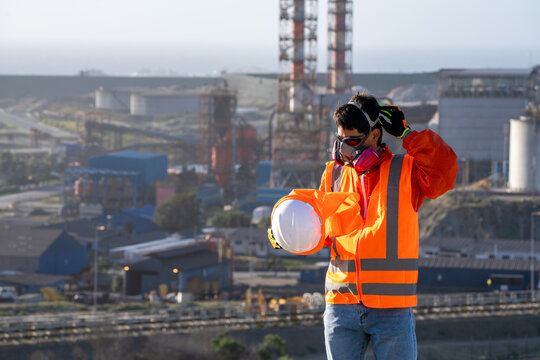Industrial worker in a gas mask, adjusting protective gear and holding a hard hat at a factory