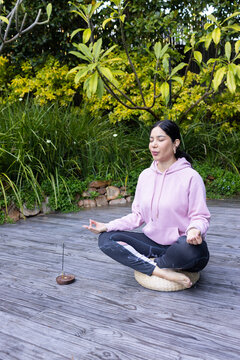 Asian woman sitting cross-legged on woven cushion meditating near incense burner on garden deck