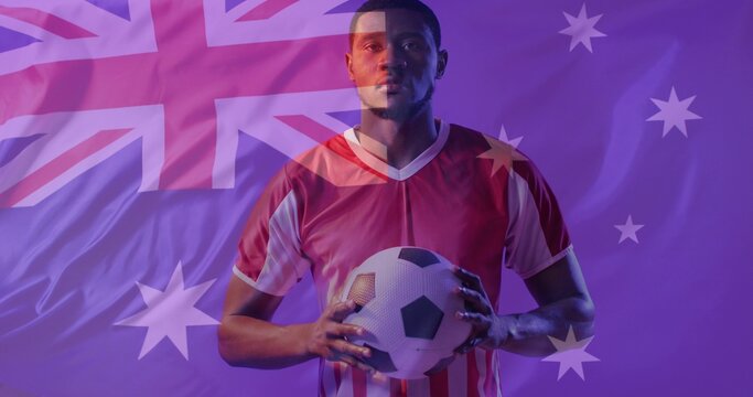 Male soccer player in red and white jersey holding ball facing purple lit Australian flag backdrop
