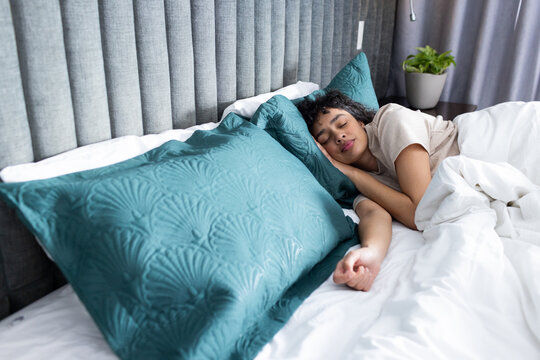 African American woman sleeping in bed with soft bedding and large teal pillows near grey headboard