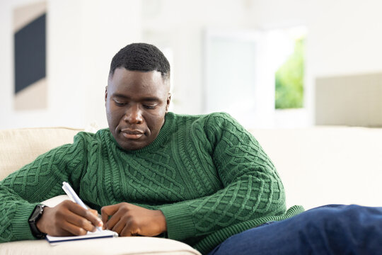 African American man in green cable-knit sweater sitting on beige couch and writing in notebook