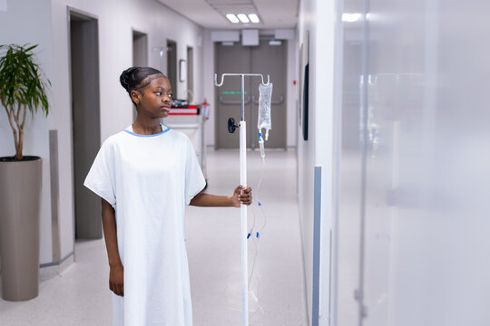 African American teenage girl in white hospital gown holding wheeled IV pole in bright hallway