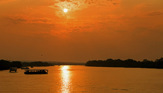 Sunset over the Zambezi River, with bright orange sky with sun reflecting in the water, with passenger tour boats. Zimbabwe, Africa