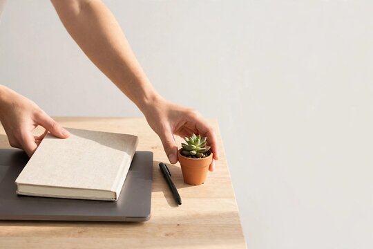 Hands tidying a desk with a blank notebook and a small plant in morning light