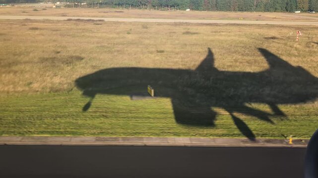 Dry grassy airfield and paved taxiway crossed by the dark moving shadow of a low-flying plane filmed from inside the aircraft to show flight paths and aviation operations for travel campaigns