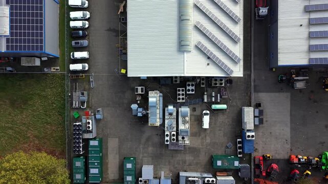 Solar panels and rows of ventilation units cover industrial rooftop beside parked cars in factory yard, revealing modern energy infrastructure with technical precision. Aerial drone top down rising