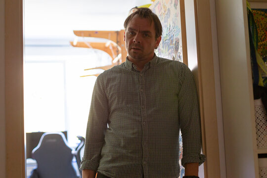 Man standing in doorway wearing checked button-up shirt, backlit by ay