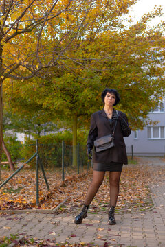 Woman in black blazer and boots standing on autumn sidewalk by fence
