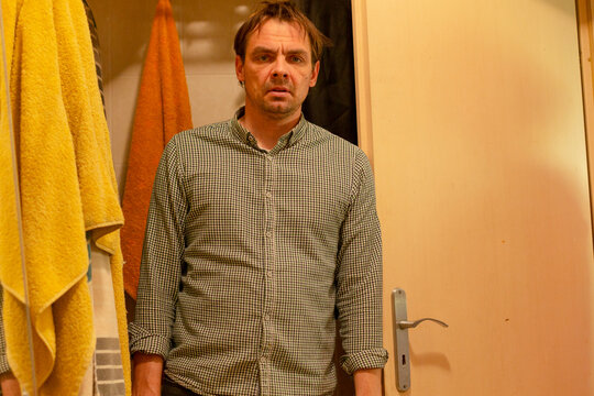 Man standing in bathroom doorway wearing checked shirt under warm lamp