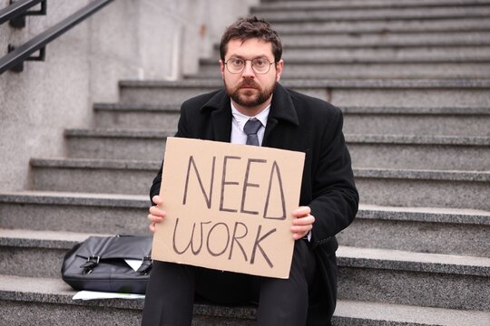 Unemployed man holding cardboard sign with words Need Work while sitting on stairs outdoors