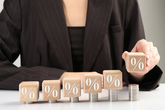 Woman placing wooden cube with percent sign onto stacked coins at table, closeup. Discount concept