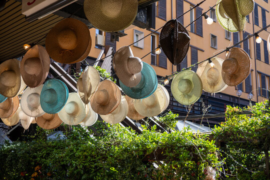 Bologna, Italy - October 14, 2026: Straw hats hanging from string lights above a street with climbing plants and an orange building in Bologna, Italy