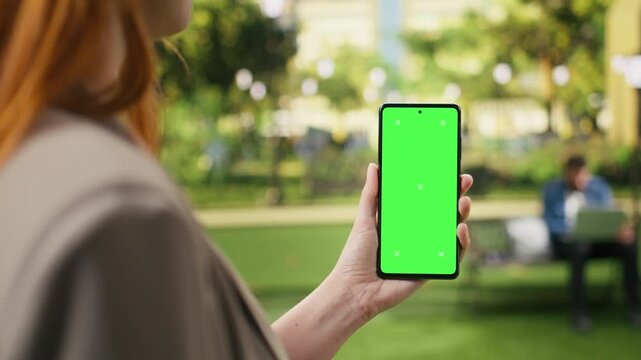 Vertical chroma key fills the smartphone in close-up as a woman works outdoors, creating a clear editable mockup for screen replacement in a modern urban park, for website content placement.
