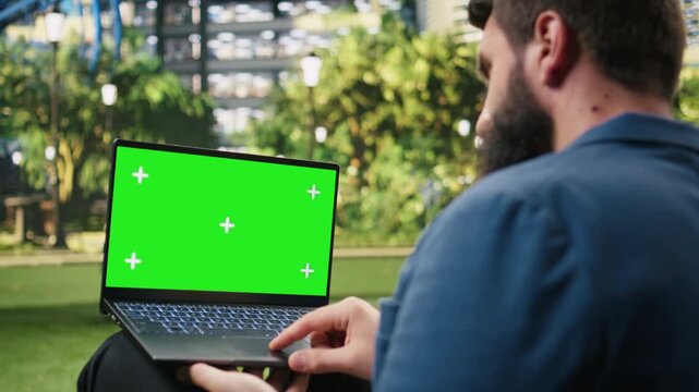 Businessman presents a laptop with a green screen display for website or advertising mockups, holding the editable widescreen against an urban park backdrop with city greenery beyond the device.