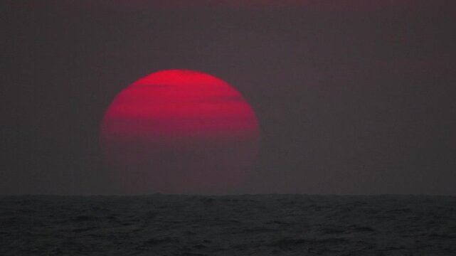 Extreme closeup of dramatic crimson red sun sphere slowly setting below dark sea horizon during evening twilight sky