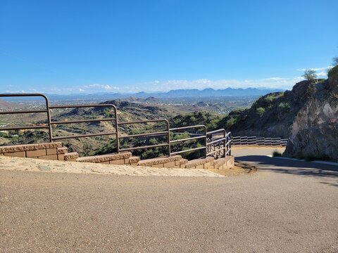 A winding paved walkway with tiered metal safety fences descends a rocky hillside overlooking a sweeping panoramic view of mountains and a flat desert valley, North Mountain park, Phoenix, Arizona 