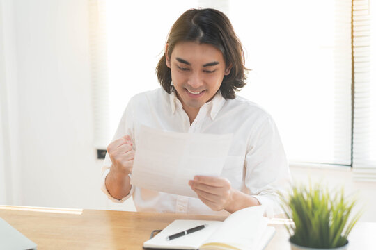 Happy young Asian man celebrating good news while reading paper document at home office desk, cheerful male student raising fist with excitement for exam results or job offer