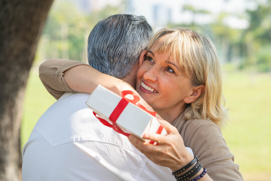 Happy senior couple embracing in sunny public park after giving gift box, mature woman hugging senior husband with love