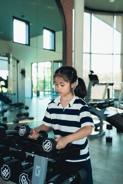Young girl picking up black dumbbell weight in modern bright gym with exercise equipment and large windows background