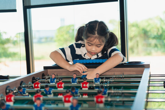 Young girl with pigtails focused on adjusting foosball player on table inside bright room with large windows and outdoor view