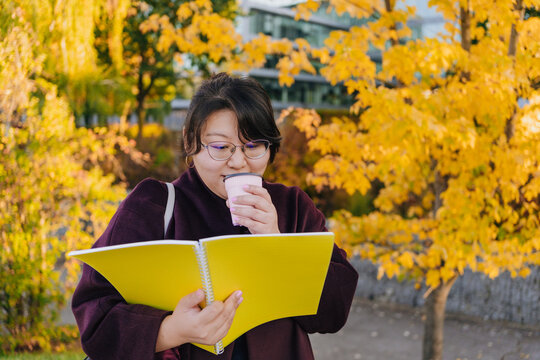 Woman reads yellow book while holding cup among autumn trees