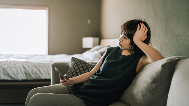 ソファでスマートフォンを使いながら考えごとをする女性