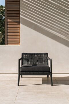 Black woven lounge chair on a warm modern exterior terrace with wood shutter and angled shadows