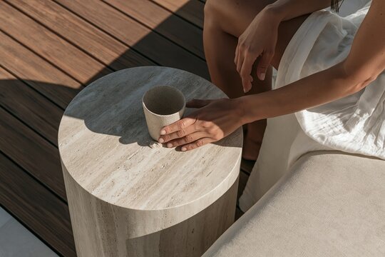 Woman in white dress holding ceramic coffee cup beside a travertine table on a warm modern terrace