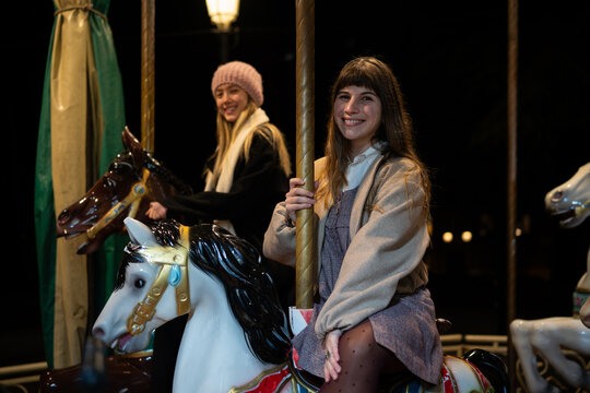 Two white women smiling on merry-go-round carousel horses at night