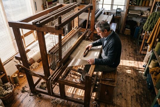 Senior male artisan weaving fabric on a traditional wooden floor loom in a sunlit workshop, carefully holding a shuttle over the threads