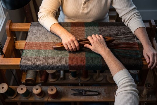 Two people passing a wooden weaving shuttle over a textured wool textile on a traditional handloom