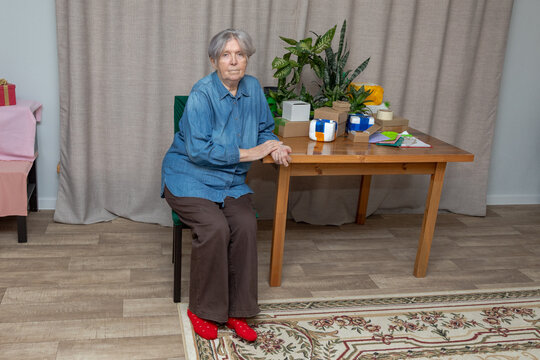 Elderly woman seated beside a table with parcels and gift boxes. Grandmother at home with packages and houseplants. Gift wrapping and holiday preparation. Family celebration and caregiving