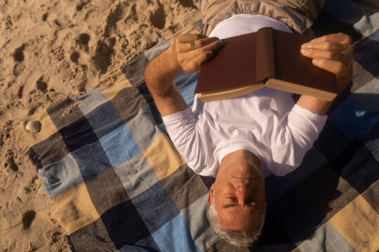 Senior man reading a book while relaxing on picnic blanket at beach