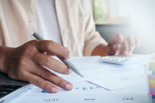 Close up of businessman hands holding pen and using calculator to analyze financial report and data document on desk in modern office, concept of accounting and audit.