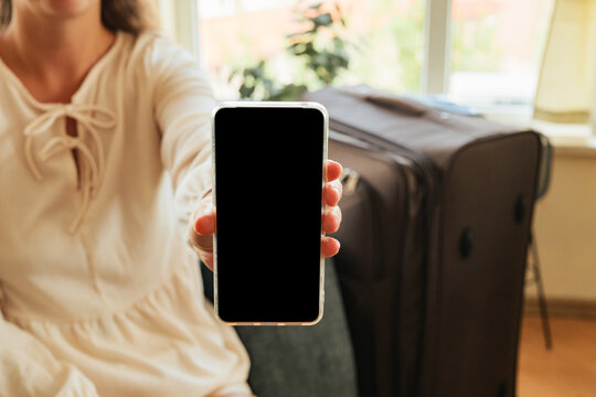 Woman showing a smartphone with a black display screen to the camera against a travel suitcase during trip preparation.