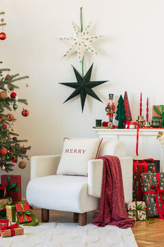 Cozy living room interior decorated for Christmas with a comfortable white armchair, glowing tree, wrapped presents, candles, and hanging paper stars