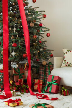 A beautifully decorated Christmas tree stands next to a sofa, surrounded by wrapped gifts and long red ribbons on the carpet, creating a warm holiday mood