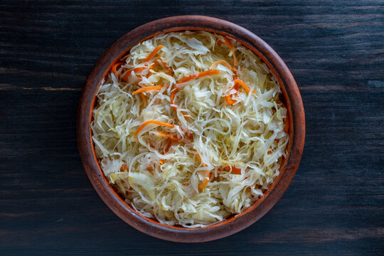 Sauerkraut with carrots in ceramic bowl on wooden background top view. Traditional fermented sauerkraut mixed with shredded carrots in rustic clay bowl on dark wooden table, flat lay