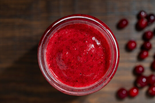 Red berry smoothie or jam in glass jar on wooden background, closeup. Healthy red cranberry smoothie or fruit puree in glass jar, shot from top view on dark wooden table with scattered berries