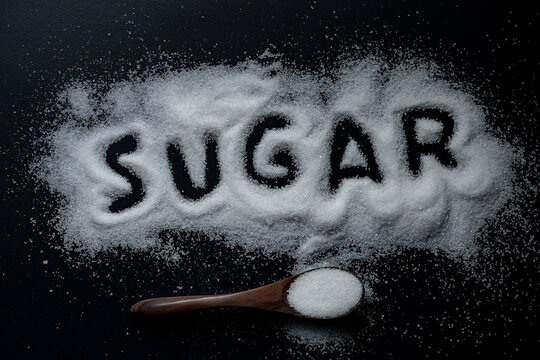 Word sugar written in white granulated sugar with wooden spoon on black background, close up, top view