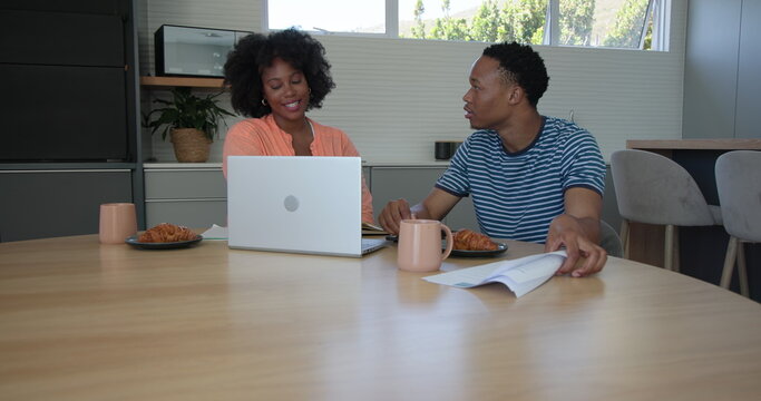 Young couple reading book and discussing notes at kitchen table with laptop