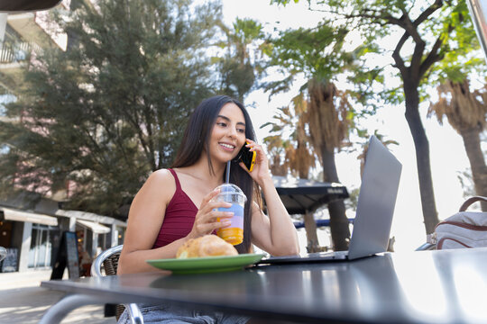Smiling woman talks on smartphone while having breakfast outdoors