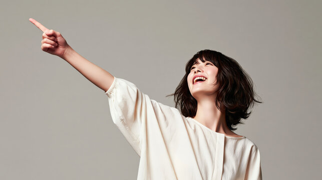 Woman pointing upward, smiling in white blouse against plain gray background, expressing joy and confidence in studio portrait